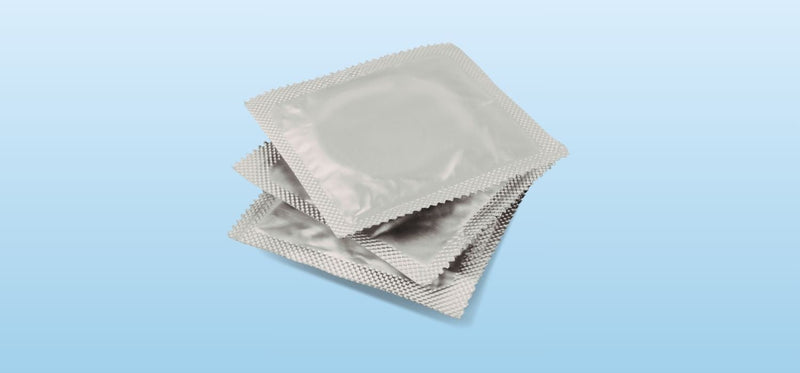A photograph of three square, silver foil condom wrappers stacked on top of each other against a light blue gradient background.