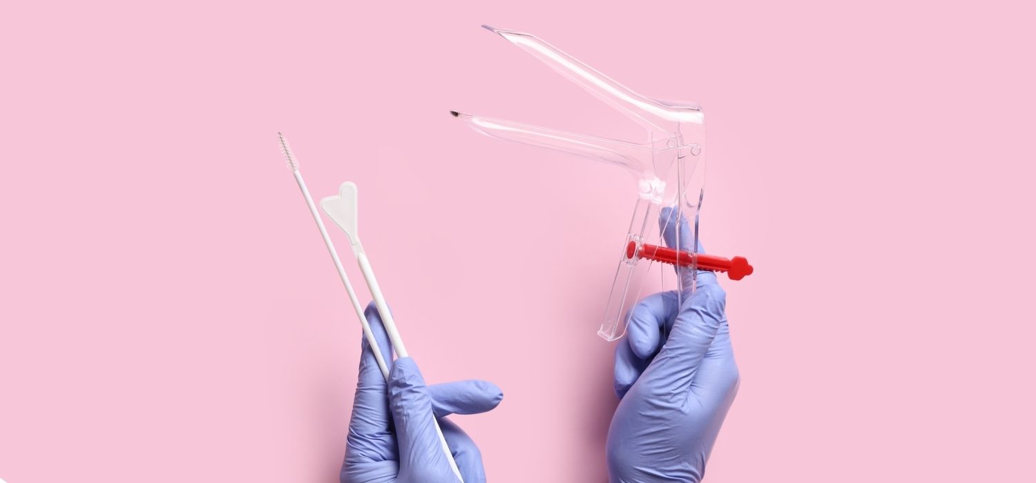 A top-down photograph against a pink background of hands in blue medical gloves holding a clear plastic speculum and two white cervical sampling swabs.