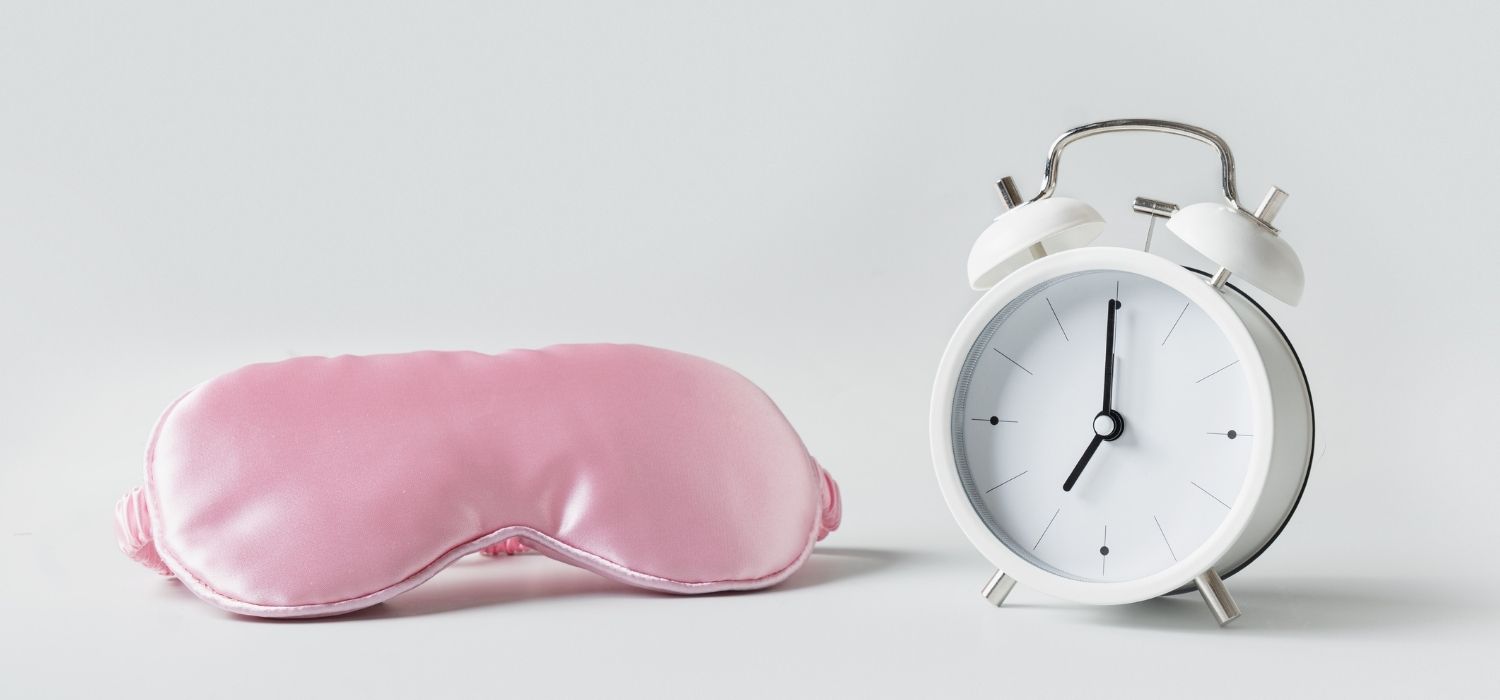 A pink sleep mask and a white alarm clock on a plain gray background.