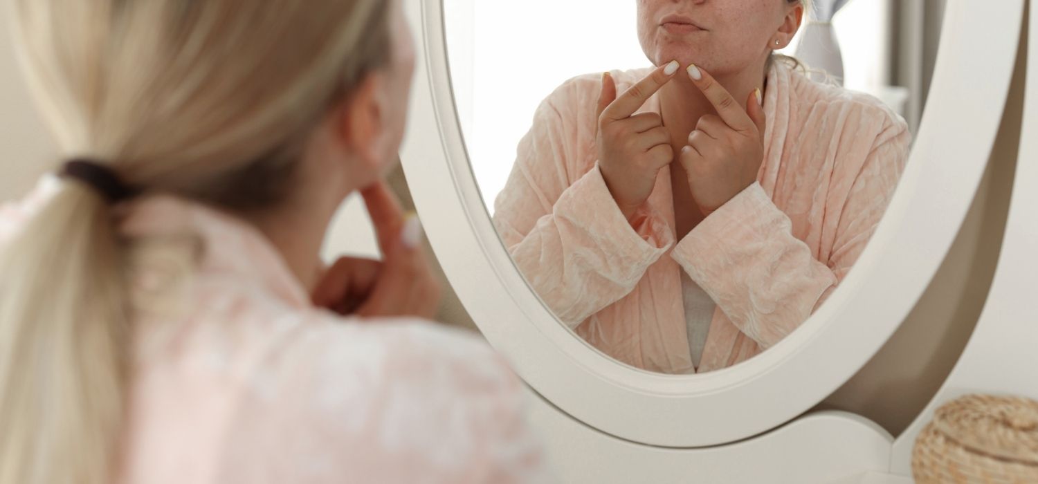 An image of a woman looking in the mirror and picking on a pimple on her face.