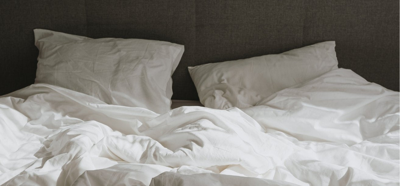 An unmade bed with rumpled white sheets, a duvet, and two pillows against a dark gray headboard.