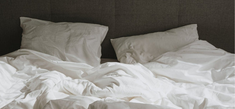 An unmade bed with rumpled white sheets, a duvet, and two pillows against a dark gray headboard.