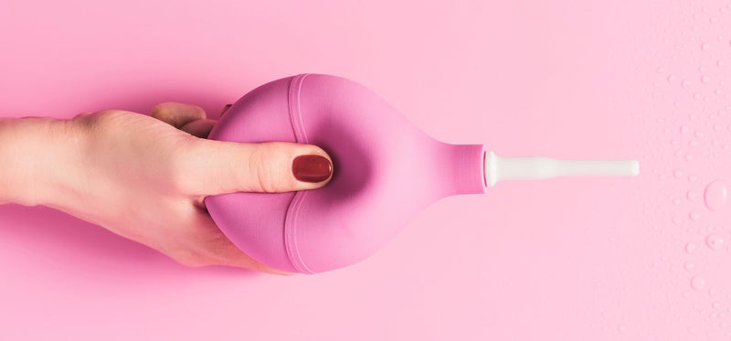 A close up of a woman's manicured hand, squeezing a pink douche, against a pink background.