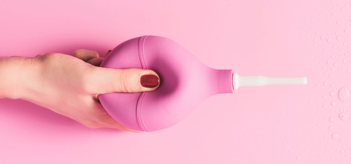 A close up of a woman's manicured hand, squeezing a pink douche, against a pink background.