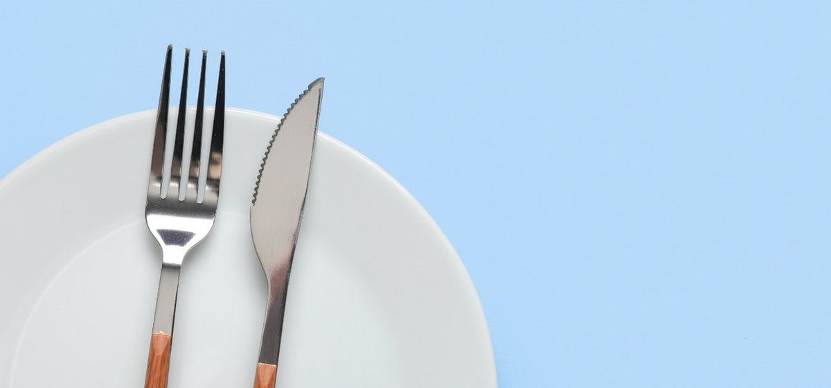A silver fork and serrated knife resting side-by-side on a white plate against a light blue background.