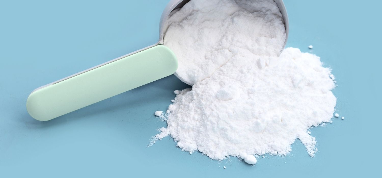 A close up photo of a tilted measuring spoon with baking soda powder spilling from one of its sides on a light blue background.