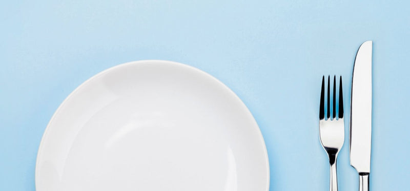 A minimalist, top-down photo of a white ceramic plate with a silver fork and knife placed neatly to the right, all set against a solid light-blue background.