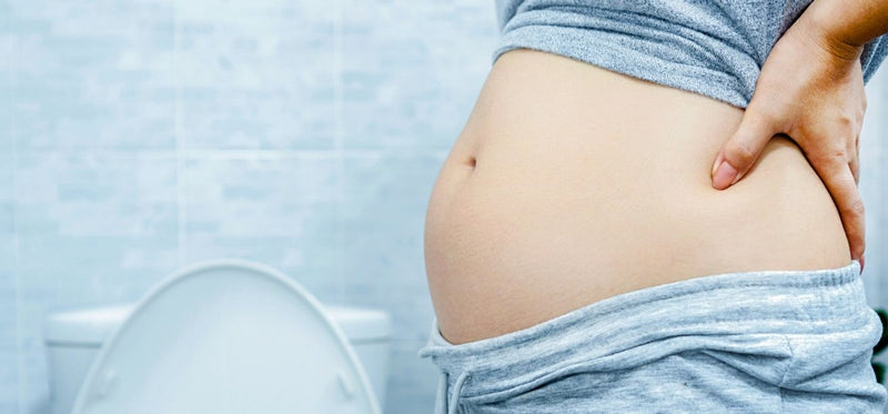 Woman in gray loungewear holding her bloated abdomen in a bathroom.
