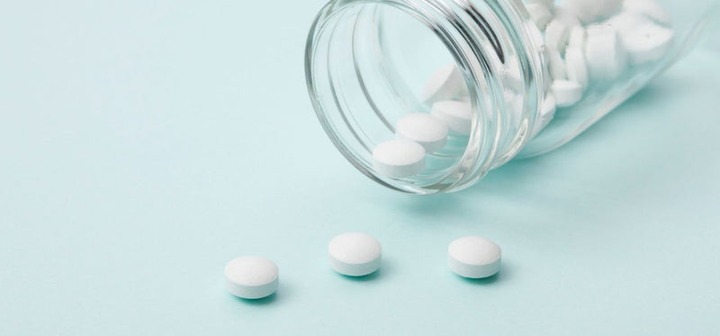 White medical pills spilling out of a clear glass bottle onto a blue background.