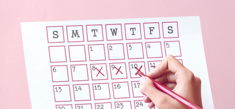 A close-up of a hand using a red pen to mark red "X"s over dates on a paper calendar, set against a solid pink background.