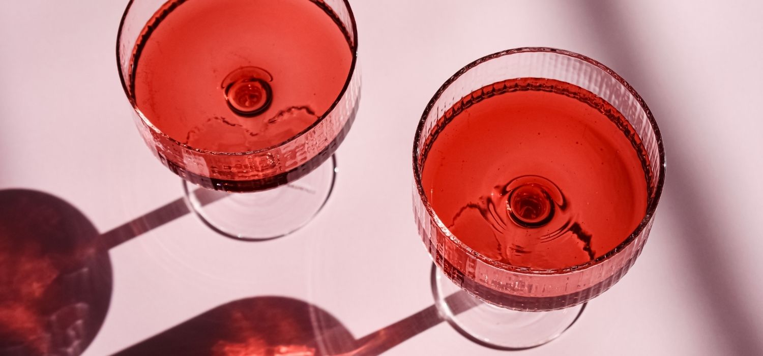 A close up image of two cocktail glasses with red liquid inside, against a pink background.
