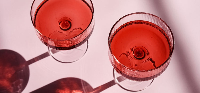 A close up image of two cocktail glasses with red liquid inside, against a pink background.