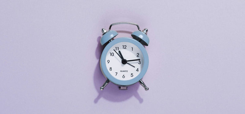 An image of an old-fashioned blue Quartz alarm clock pinned to a light purple background.
