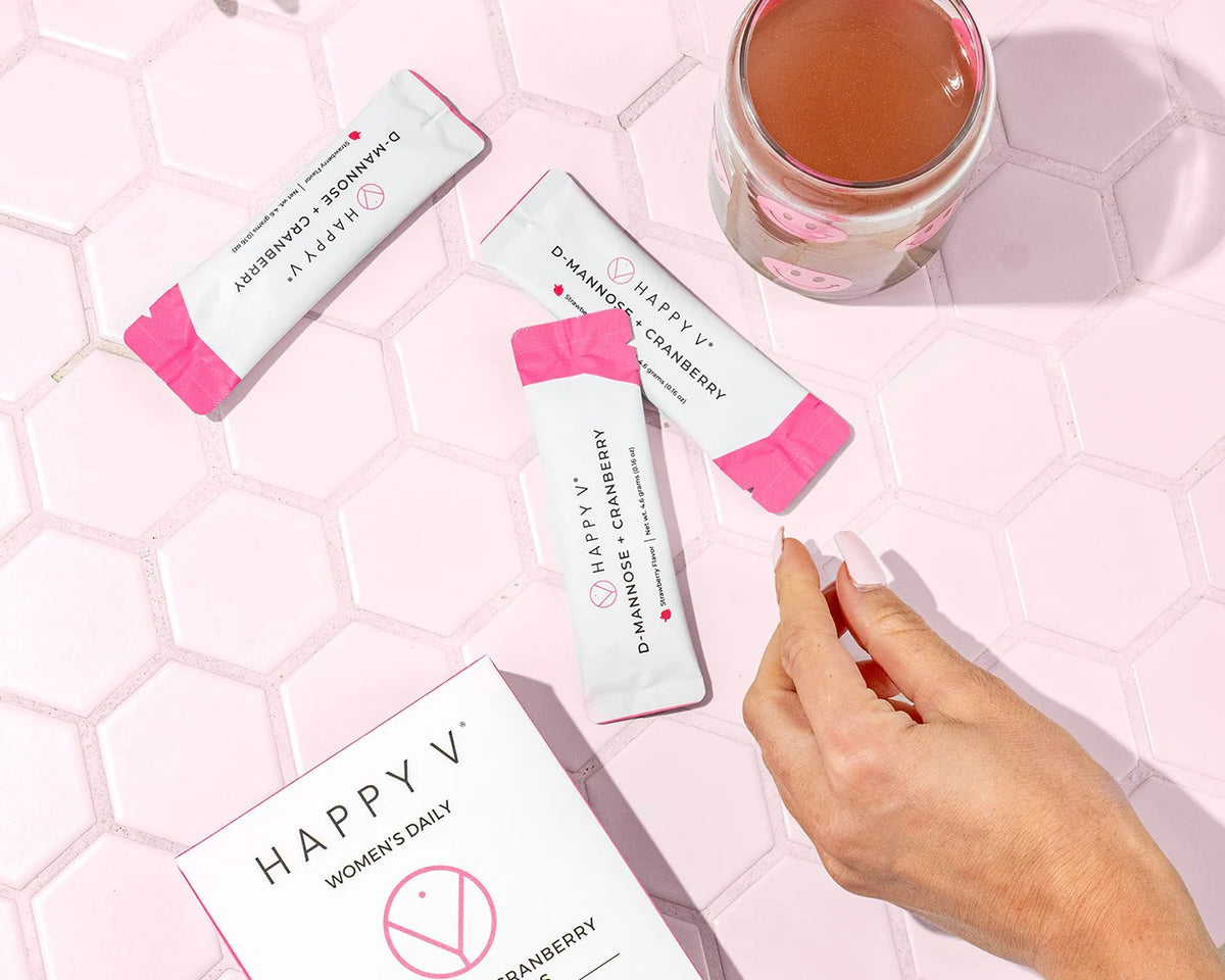 A box of Happy V's D-Mannose and Cranberry stick packs with some stick packs laying next to a glass of diluted Cranberry and D-mannose supplements.