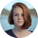 Woman with short hair smiling, holding a Happy V feminine wellness supplement bottle outdoors.