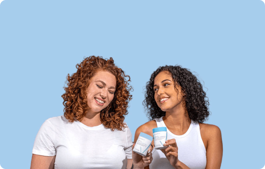 Two women smiling and holding skincare products against a light blue background.