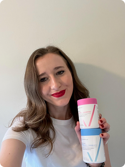 Woman holding two colorful supplement containers from Happy V against a plain wall.