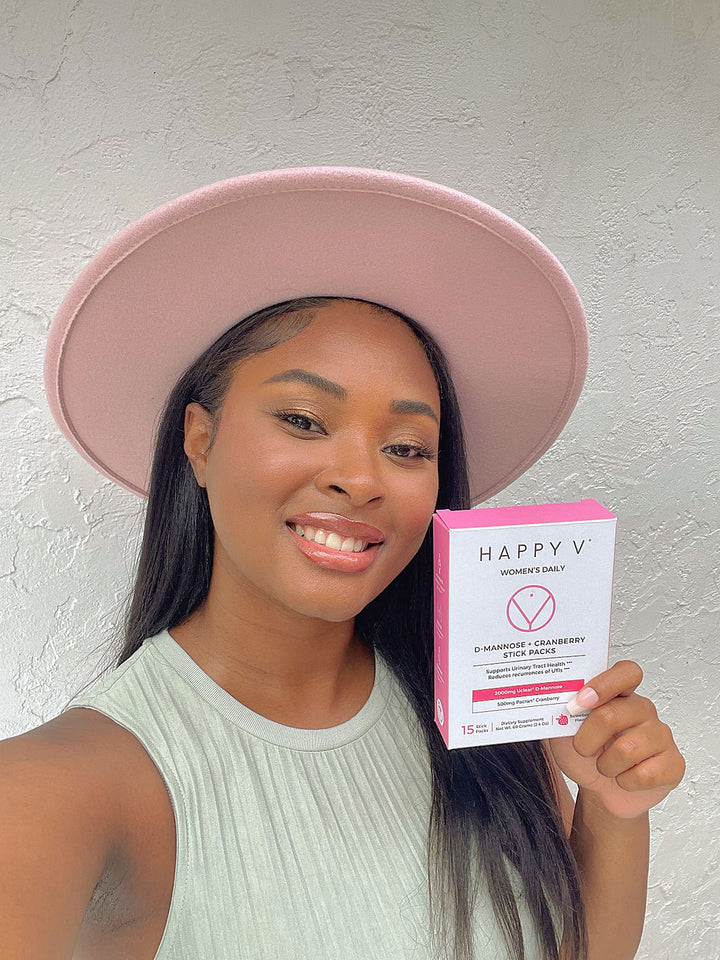 A smiling girl in a pink hat holding a box of Happy V D-mannose and Cranberry Stick Packs