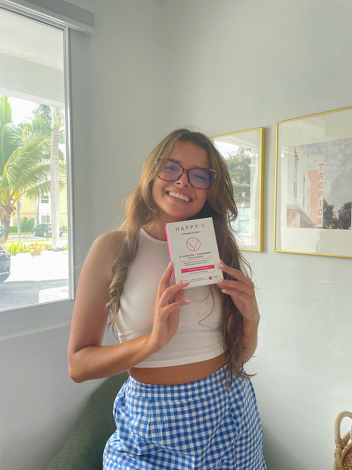 A smiling girl holding a box of Happy V D-mannose and Cranberry Stick Packs next to her face
