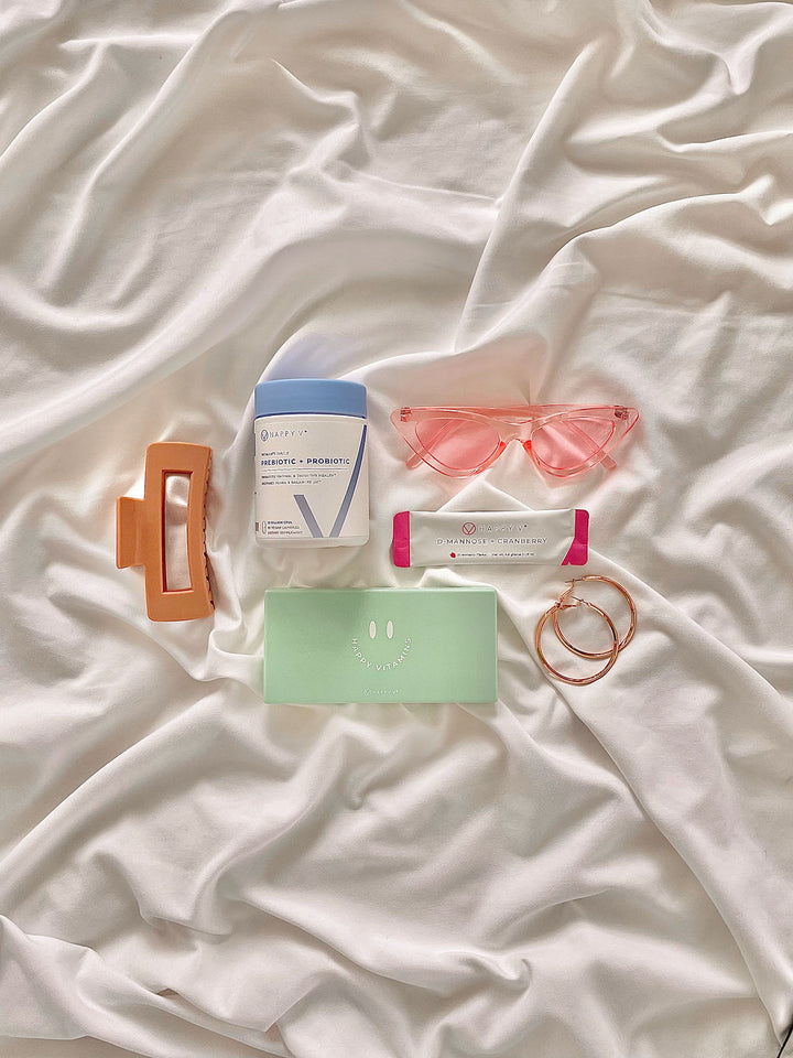 Happy V Prebiotic and Probiotic Supplements bottle laid next to a Happy V D-mannose and Cranberry Stick Pack, a hair clip, two hari ties, and a pair of pink sunglasses