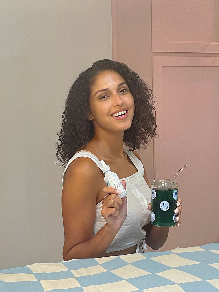 A smiling girl with curly hair holding a Happy V glass full of Happy V Liquid Chlorophyll in one hand and a bottle of Happy V Liquid Chlorophyll in the other hand