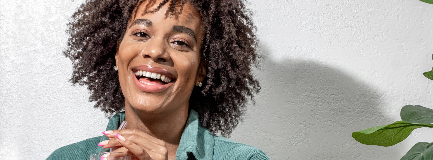 A close up of a smiling woman with curly hair drinking with a glass straw from a glass blog banner image