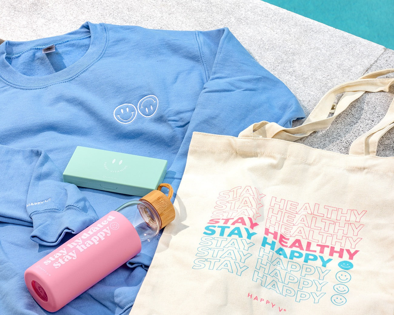 Blue sweatshirt, pink water bottle, green Happy V supplement box, and tote bag on a table.