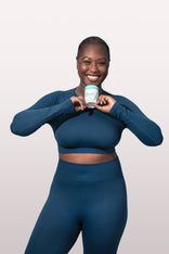 Smiling woman in activewear holding a skincare product against a light background.