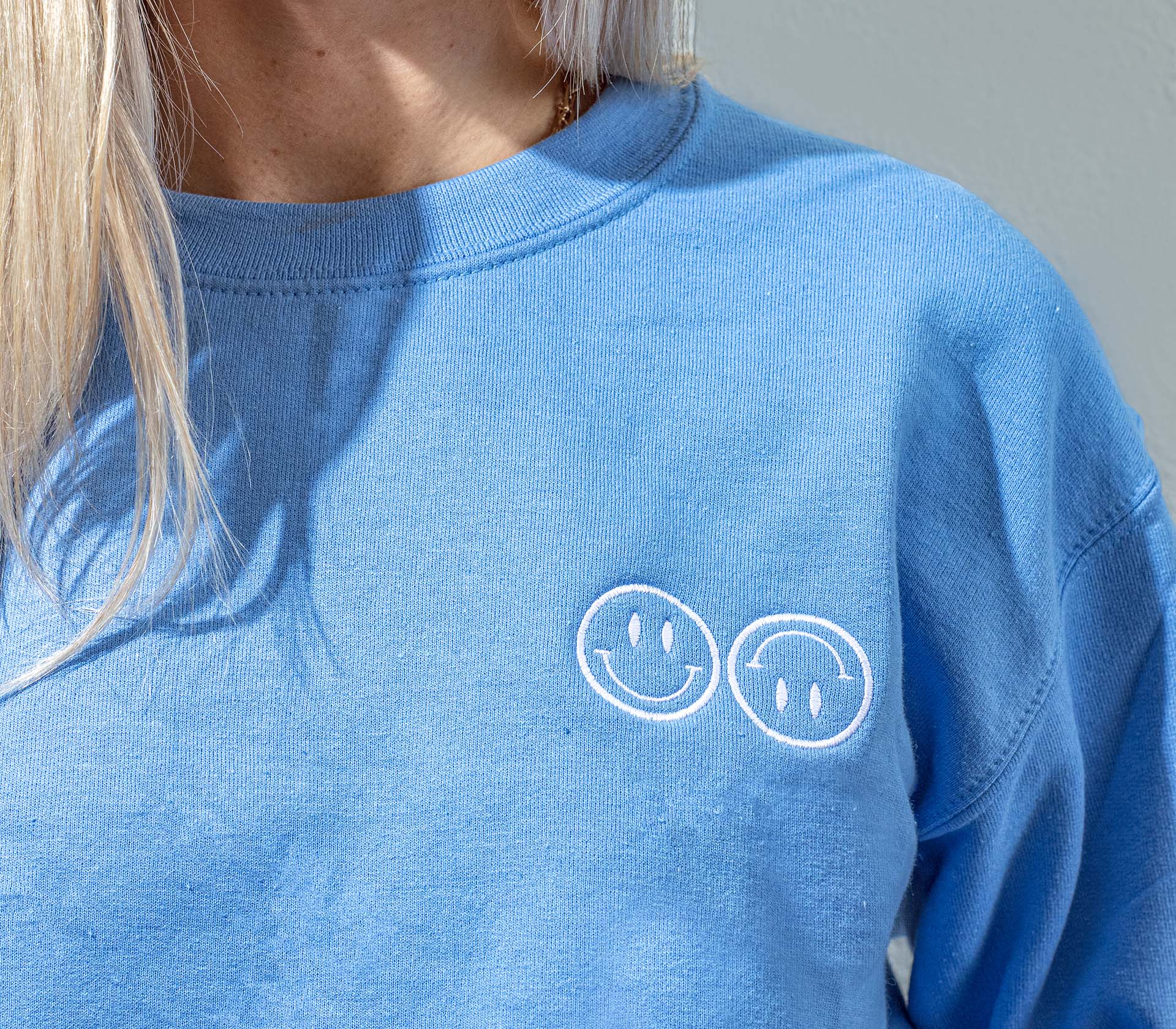 A close up of a woman wearing a blue Happy V Crewneck Sweatshirt, displaying the Happy V logo.