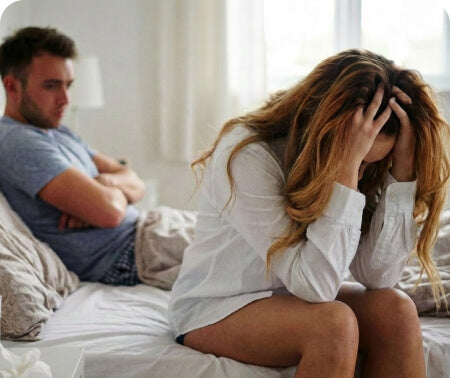 A woman sits on a bed with her head in her hands while a man sits nearby, both appearing distant.