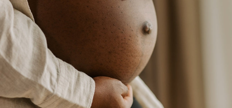 Close-up of a person of color, holding their bare pregnant belly from underneath.