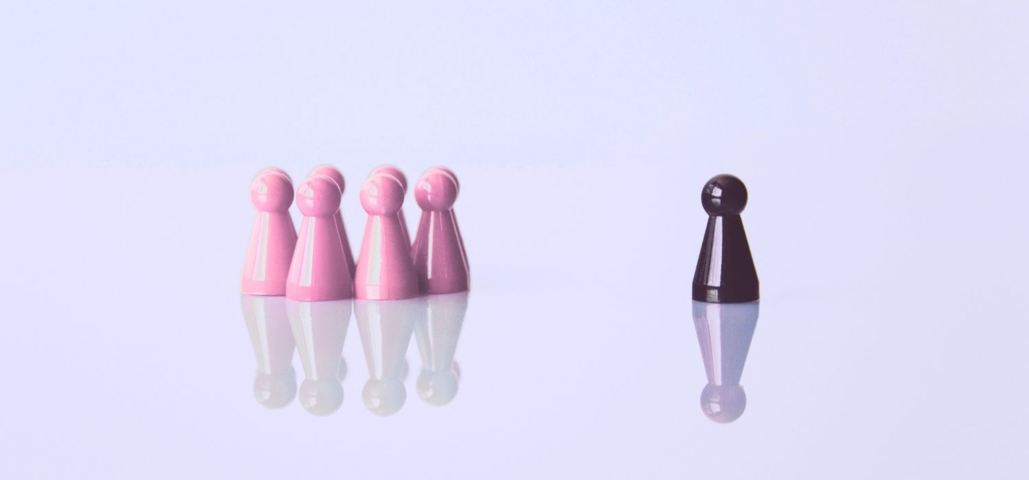 A group of pink board game pawns standing together, separated from a single black pawn on a reflective white surface.