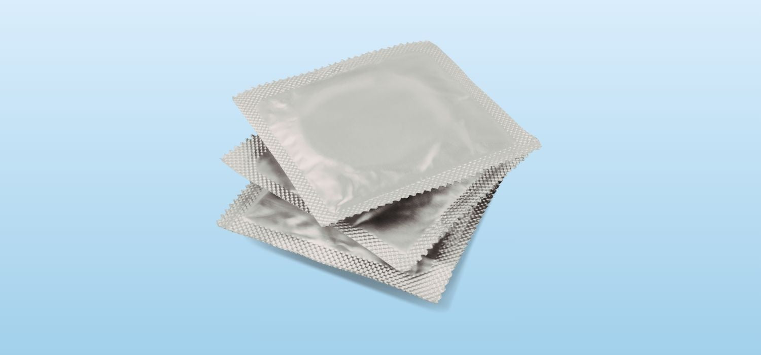 A photograph of three square, silver foil condom wrappers stacked on top of each other against a light blue gradient background.