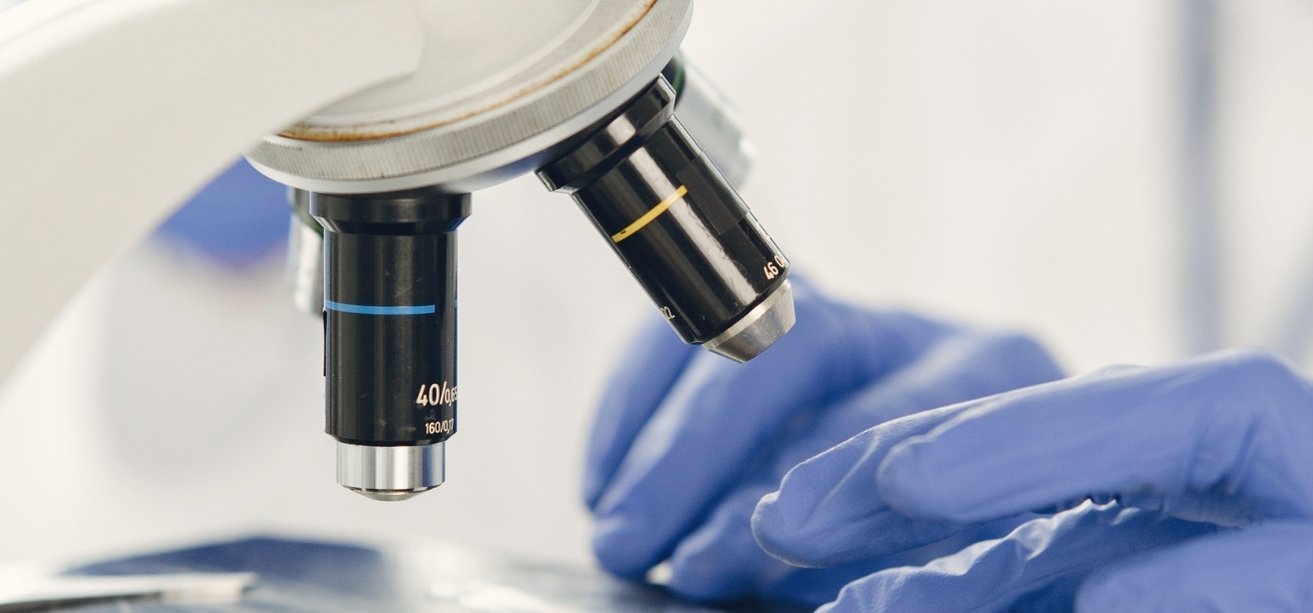 A close-up of microscope objective lenses with a blurred background of a researcher wearing purple gloves working in a lab.