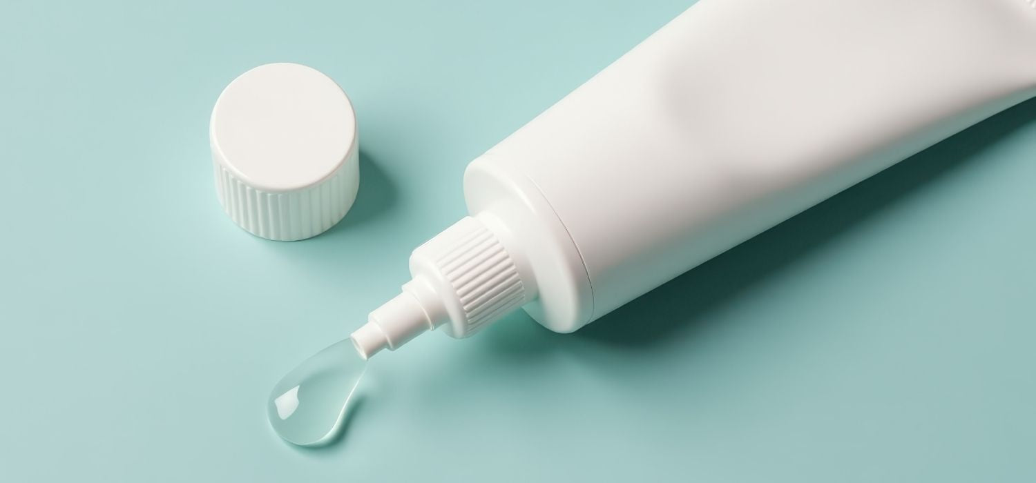 Close-up of a white medicinal tube dispensing a drop of clear gel onto a teal background.