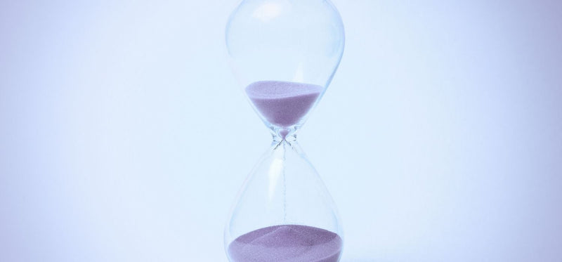 Descriptive: A clean, minimalist photograph of a glass hourglass with light purple sand flowing from the top bulb to the bottom, set against a soft blue and white background.
