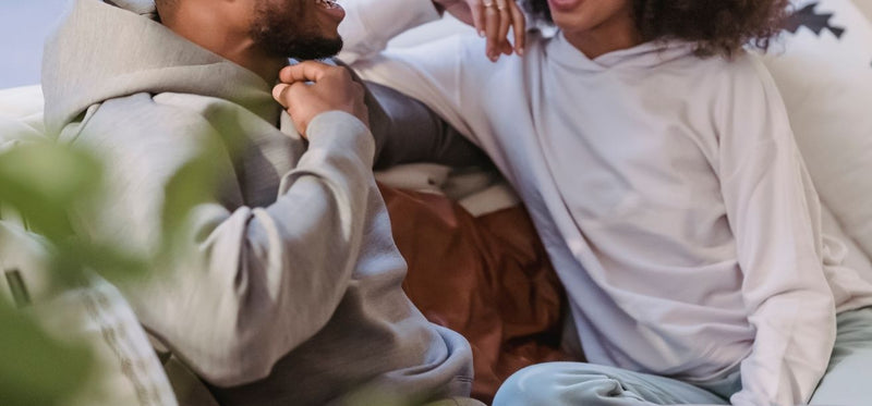 Close-up of a couple relaxing together on a couch, wearing comfortable casual hoodies while smiling and talking.