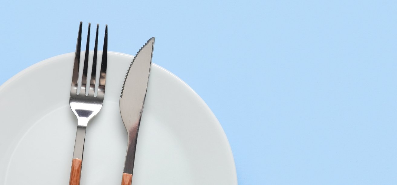 A silver fork and serrated knife resting side-by-side on a white plate against a light blue background.