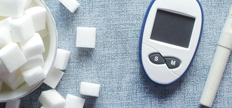 A blood glucose monitor next to a pile of white sugar cubes on a textured blue surface.