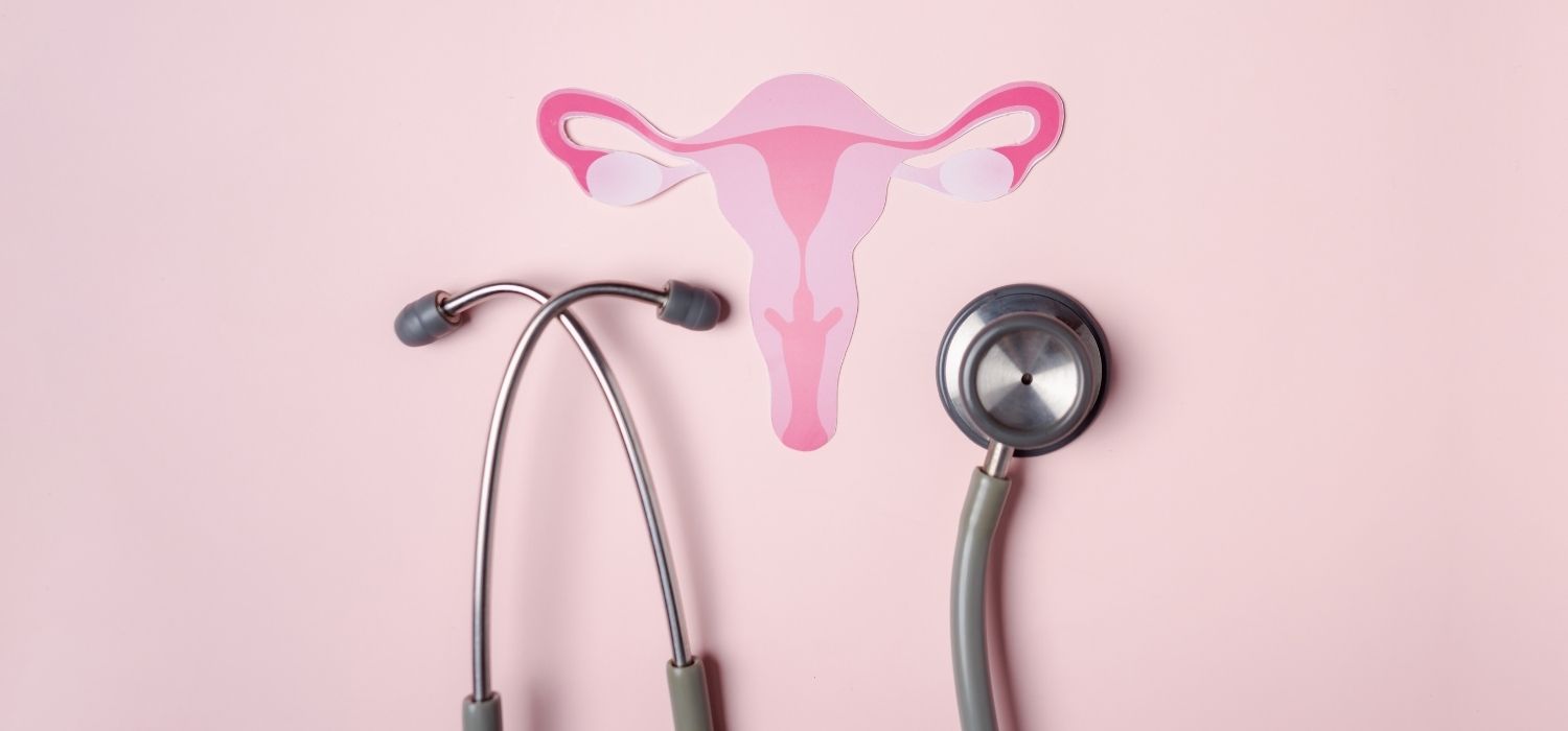 A flat-lay photograph on a pastel pink background featuring a pink paper cutout of the female reproductive system (uterus, fallopian tubes, and ovaries) positioned above a medical stethoscope.