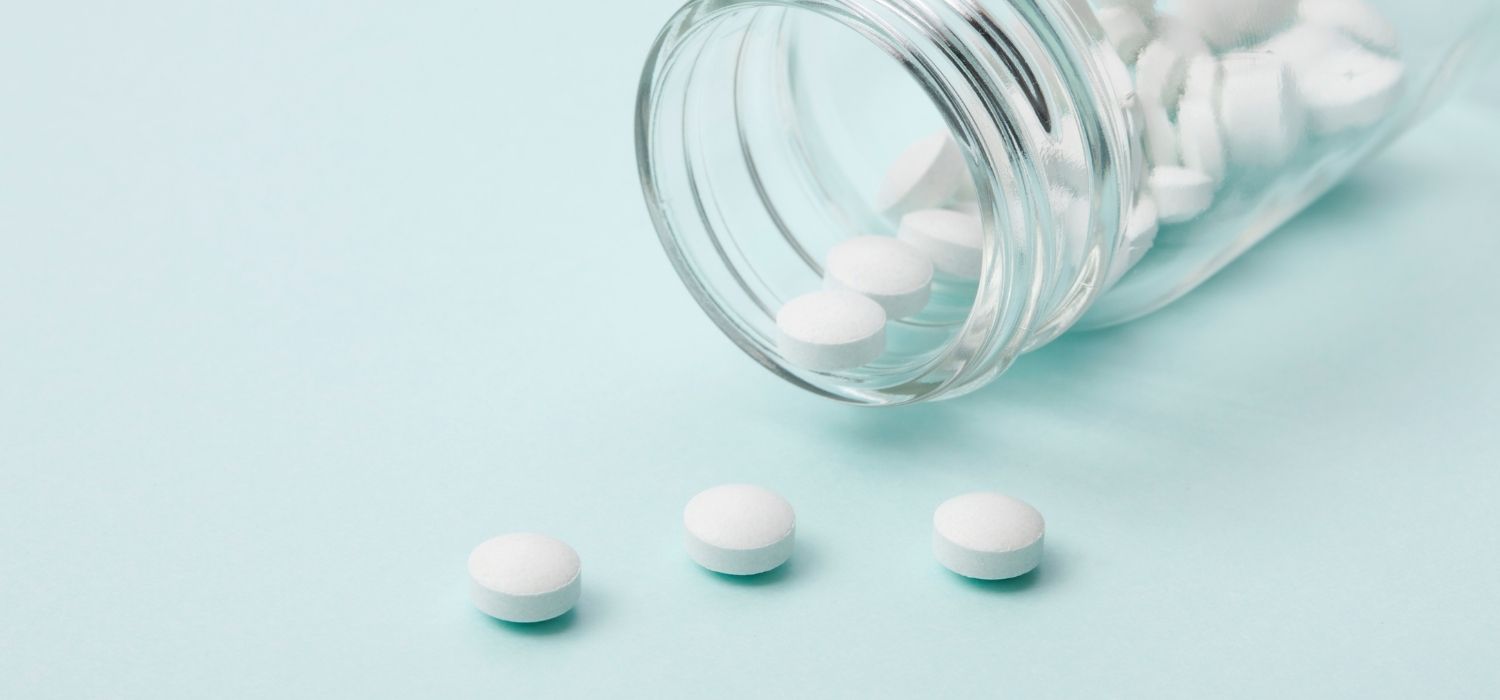 White medical pills spilling out of a clear glass bottle onto a blue background.