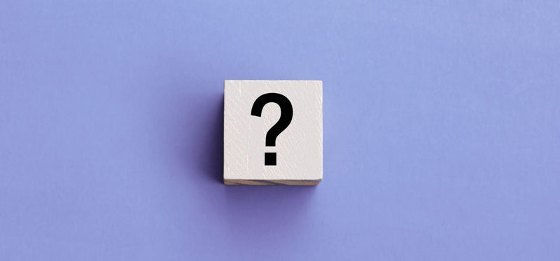 A minimalistic photograph featuring a single white wooden block centered on a solid purple background. The front face of the block is printed with a large, black question mark.