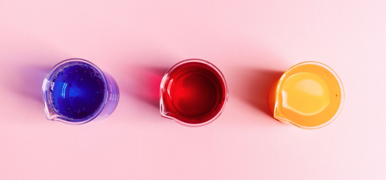 A top-down view of three glass beakers arranged in a horizontal line on a light pink surface. Each beaker contains a different brightly colored liquid: deep blue on the left, ruby red in the center, and bright orange-yellow on the right.