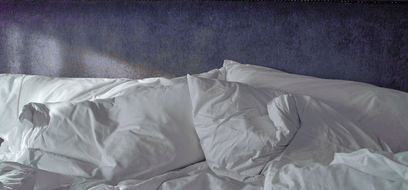 Messy white pillows and rumpled sheets on a bed with a dark purple velvet headboard, suggesting a restless night.