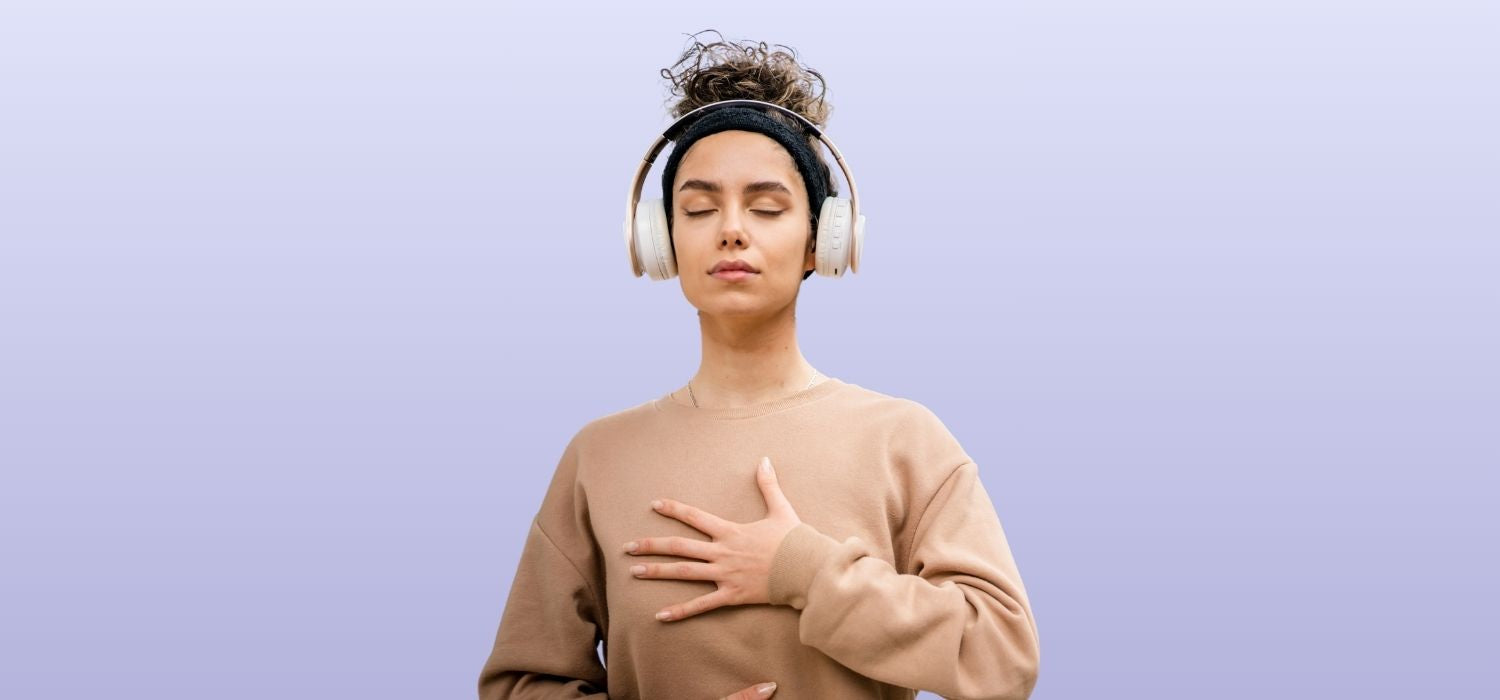A young woman with curly hair pulled up in a headband stands against a purple background with her eyes closed in a meditative pose.