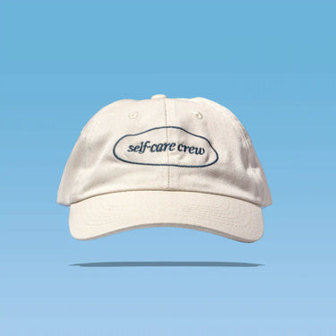 Beige cap with “self-care crew” text on blue, reflecting Happy V’s wellness style.