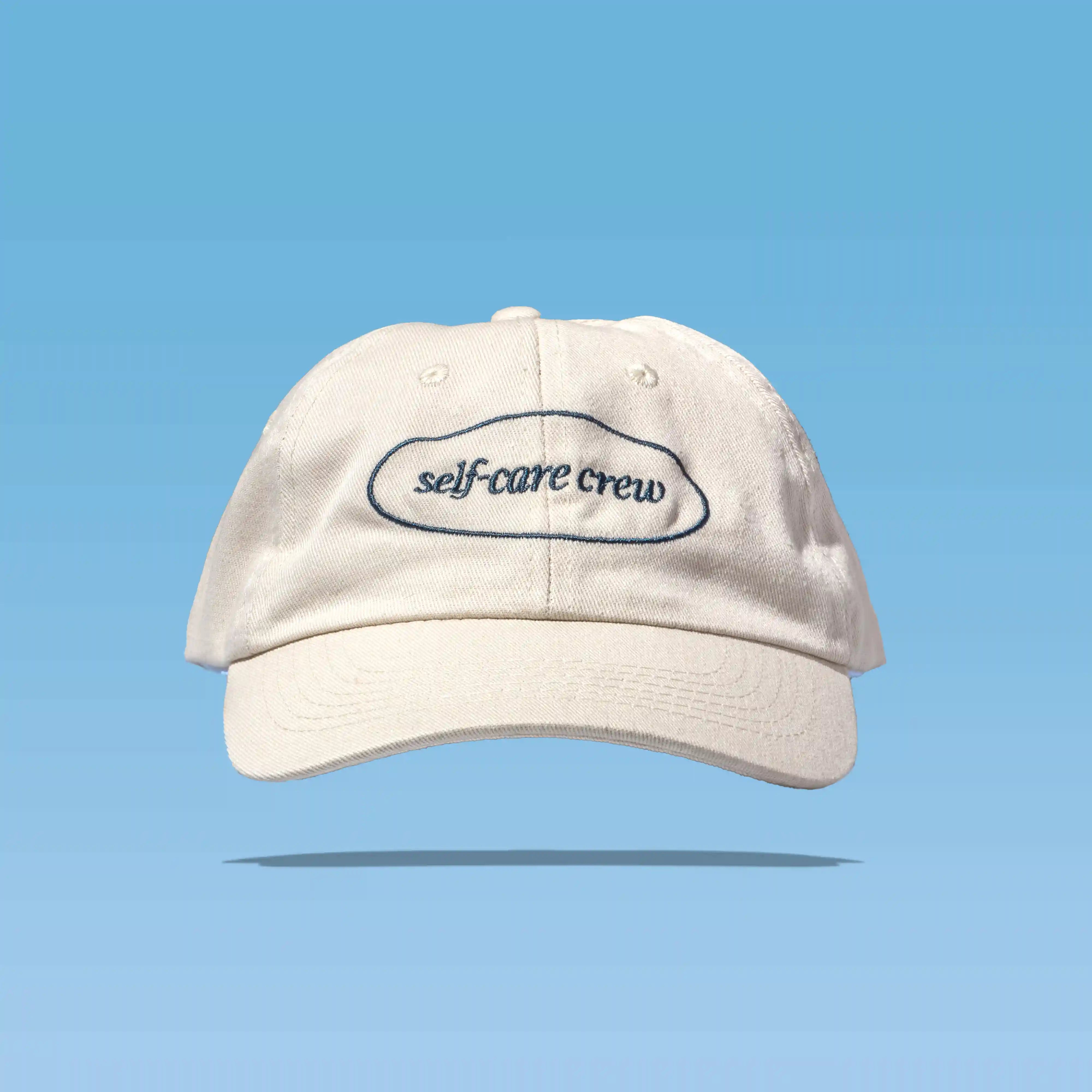 Beige cap with “self-care crew” text on blue, reflecting Happy V’s wellness style.