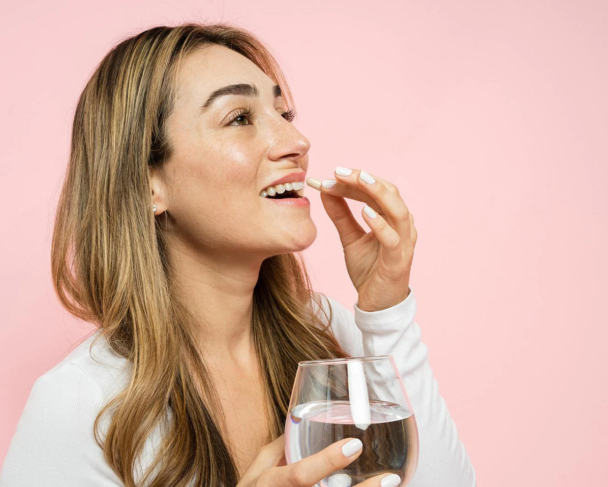 A smiling woman, holding a glass of water and about to drink a vitamin pill.