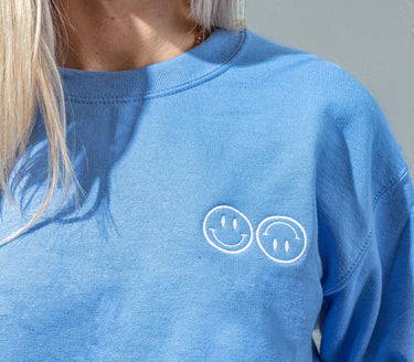 A close up of a woman wearing a blue Happy V Crewneck Sweatshirt, displaying the Happy V logo.